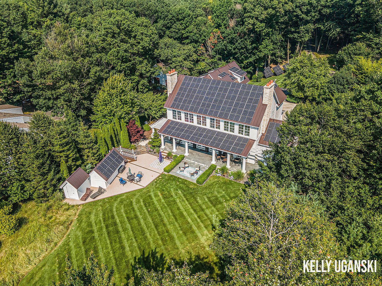 This aerial shot showcases a grand residence nestled among lush greenery. The house features a roof covered in solar panels, a spacious patio with outdoor seating, and a meticulously manicured lawn with striped patterns. The overall impression is one of luxury, sustainability, and privacy.