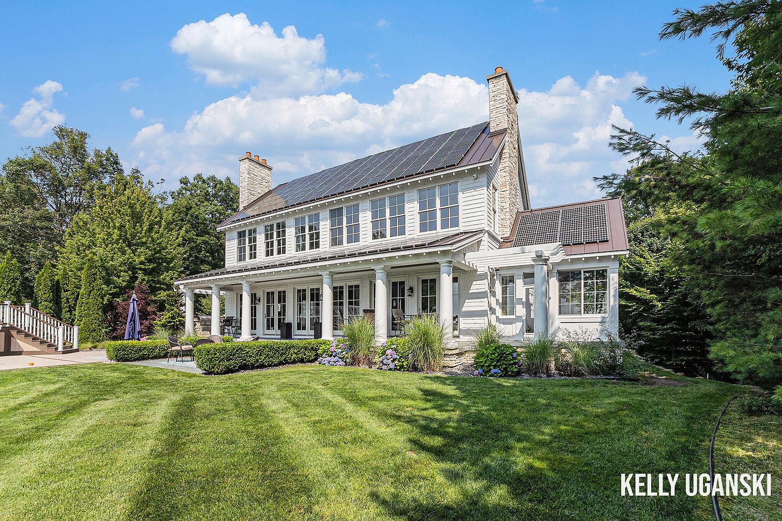 This is a front view of a large, two-story white house with a prominent porch supported by white columns. The house features multiple windows, a stone chimney, and solar panels on the roof. The well-manicured lawn and landscaping add to the property's curb appeal, creating an inviting and luxurious impression.
