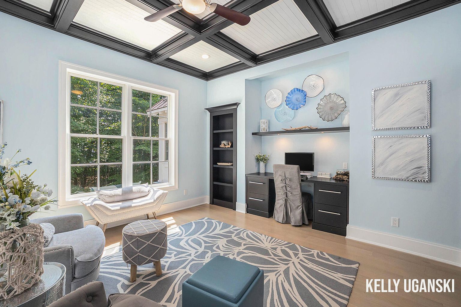 This is an interior shot of a well-lit office or study. The room features a large window, a built-in desk and shelving unit, and a coffered ceiling. The color palette is soft and calming, with light blue walls and dark wood accents, creating a sophisticated and comfortable workspace.