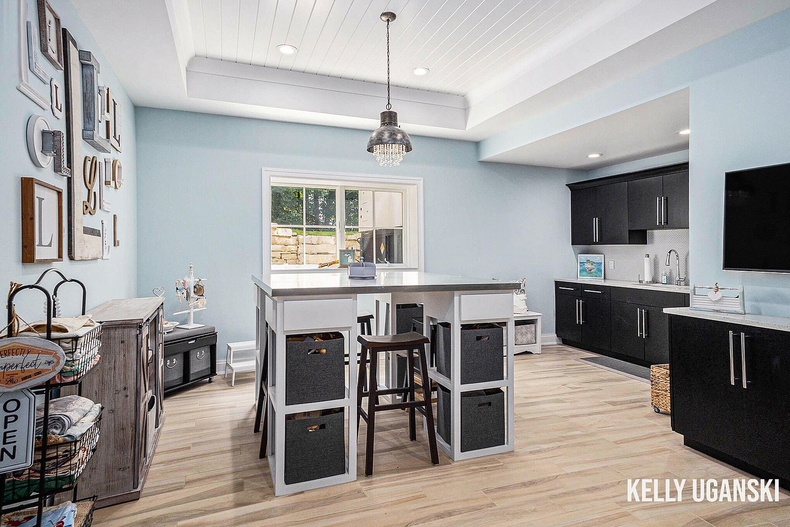 This is a well-lit basement area featuring light blue walls and light wood-look flooring. A central island with storage and seating is the focal point, complemented by dark cabinetry along the back wall with a sink and countertop area. The space is decorated with various decorative items and has a clean, organized appearance, suggesting a versatile and functional living area.