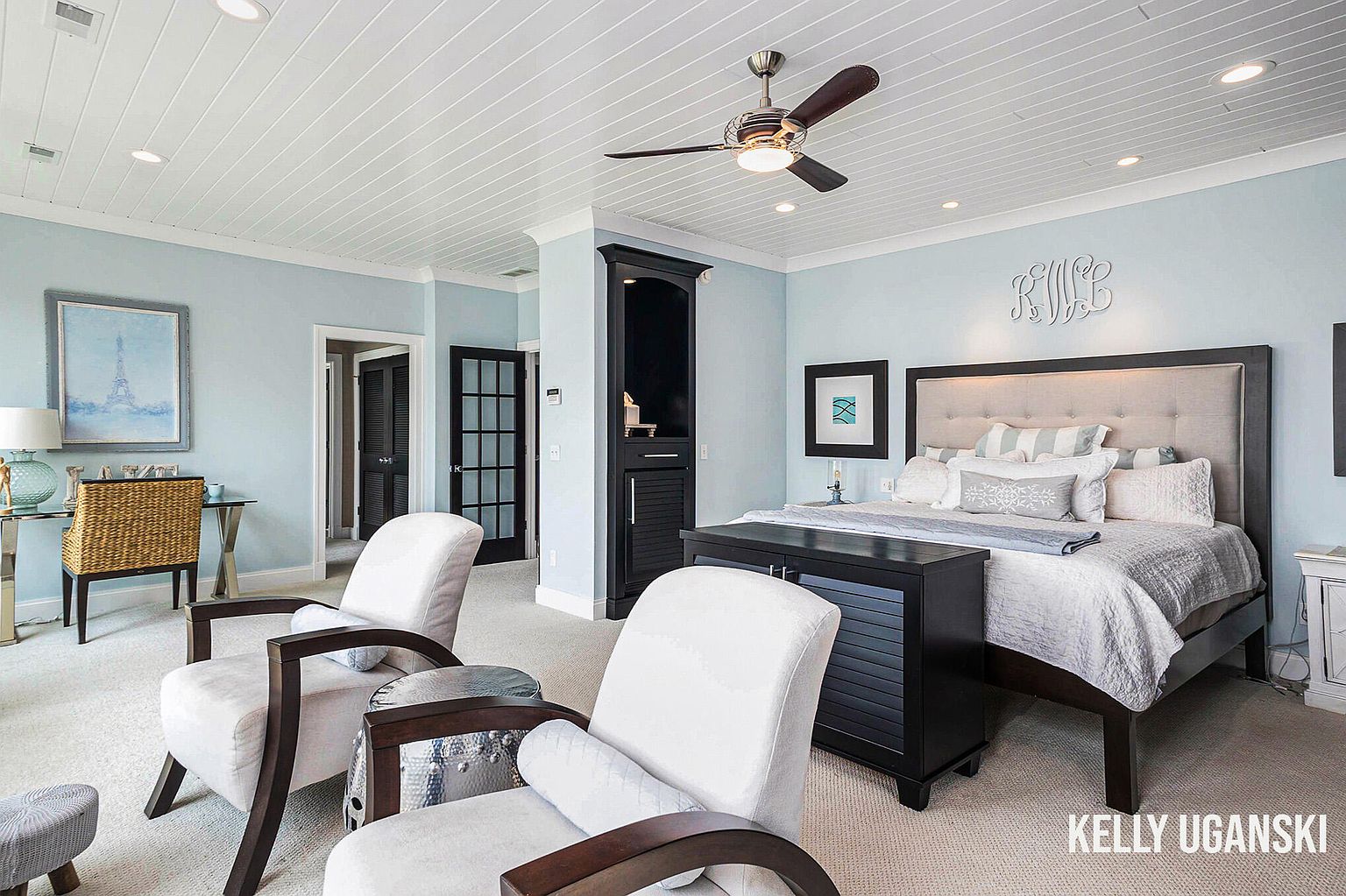 This primary bedroom features a calming blue color scheme, complemented by white trim and a coffered ceiling with a ceiling fan. The room includes two armchairs, a bed with a tufted headboard, and a dark wood dresser at the foot of the bed. A built-in cabinet and artwork add to the room's sophisticated and comfortable ambiance.