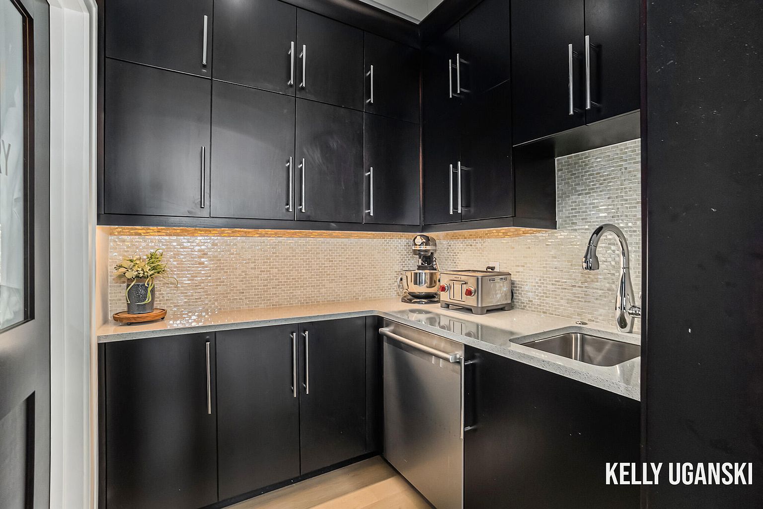 This is a modern kitchen featuring sleek, dark cabinetry with silver hardware. The countertops are a light gray, and the backsplash is a mosaic tile. Stainless steel appliances, including a dishwasher, sink, toaster, and stand mixer, are visible, adding a touch of elegance to the space.