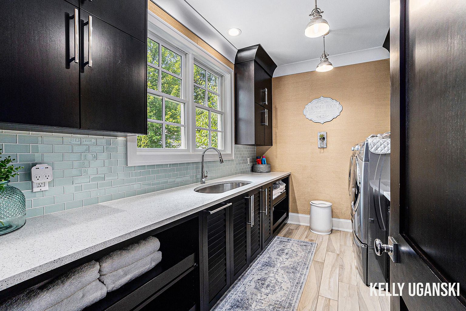 This is a well-organized laundry room featuring dark cabinetry with sleek hardware, a light-colored countertop, and a blue tile backsplash. A stainless steel sink is integrated into the countertop, and open shelving provides storage for towels. The room is illuminated by pendant lights and recessed lighting, creating a clean and functional space.