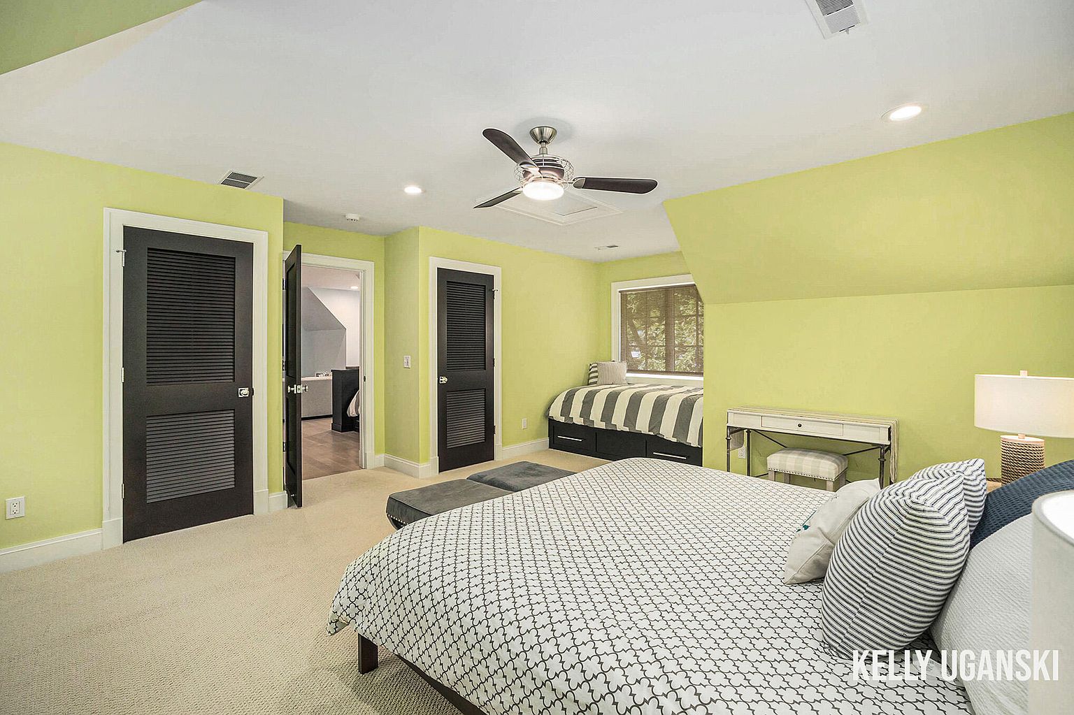 This is a bright and inviting guest bedroom featuring a light green color scheme. The room includes a bed with a patterned duvet, a small desk, and a built-in daybed by the window. Dark-colored louvered doors add a touch of contrast, and a ceiling fan provides air circulation. The overall impression is clean and comfortable.