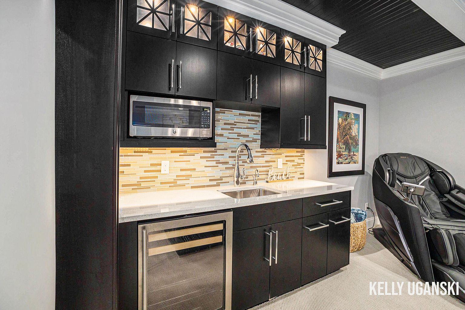 This interior shot showcases a stylish basement bar area with dark cabinetry, stainless steel appliances, and a mosaic tile backsplash. The bar features a sink, microwave, and wine refrigerator, complemented by a light countertop. A massage chair sits nearby, suggesting a relaxation zone within the basement space.