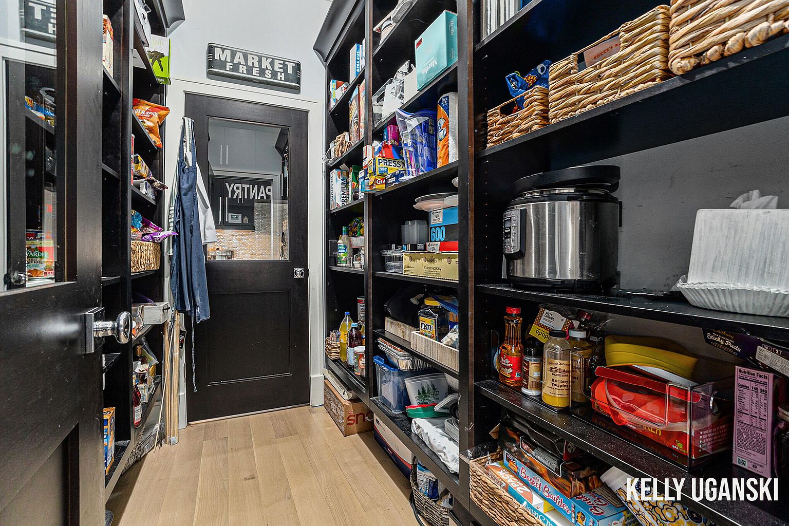 This is an interior shot of a well-organized pantry. The pantry features dark shelving units stocked with a variety of food items, containers, and appliances. A black door with a glass panel provides access to the pantry, and the light wood flooring adds warmth to the space.