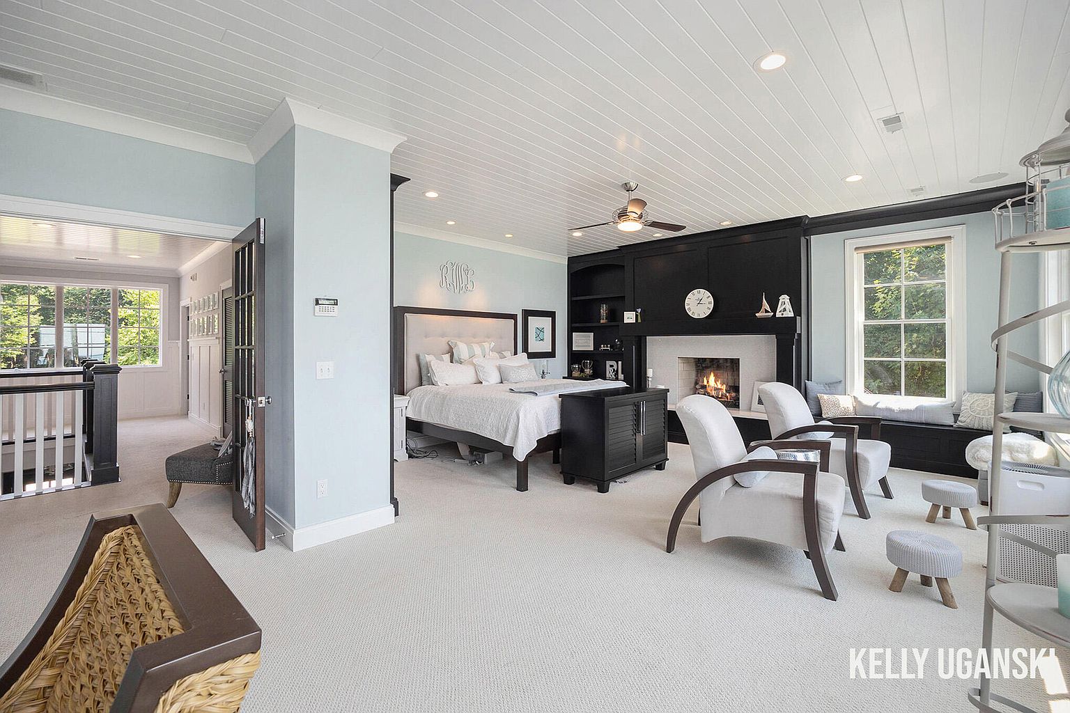 This is a spacious primary bedroom featuring a king-size bed with a tufted headboard, a fireplace with a dark wood surround, and a cozy window seat. The room is painted in light blue tones with white trim and a white plank ceiling, creating a serene and inviting atmosphere. The overall style is elegant and comfortable, perfect for relaxation.