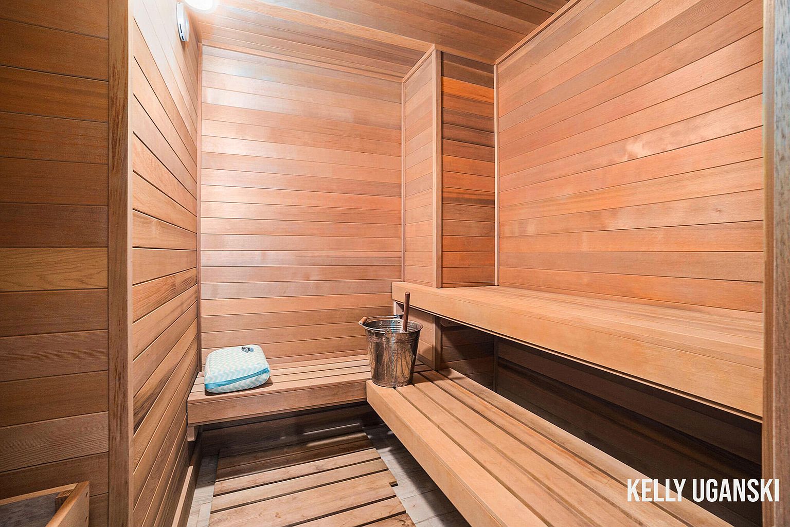 The image showcases a well-maintained sauna, featuring wooden walls, benches, and flooring. A metal bucket and a small cushion add to the sauna's functionality and comfort. The warm tones of the wood create a relaxing and inviting atmosphere, perfect for a home spa experience.