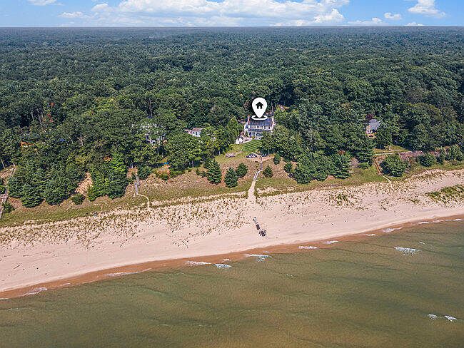 This aerial view showcases a beautiful property nestled between a lush forest and a sandy beach. The house, marked with a location pin, features a light-colored exterior and is situated on a well-maintained lawn. The scene captures the serene coastal living experience, highlighting the proximity to both nature and the waterfront.