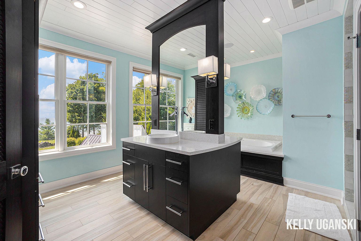 This is a primary bathroom featuring a dark wood vanity with a white countertop and a vessel sink. The walls are painted a light blue, and decorative glass plates adorn the wall near the bathtub. The flooring is light wood-look tile, and the overall style is modern and elegant.