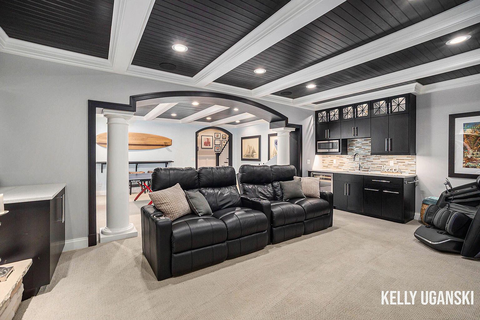 This is a well-appointed basement featuring a home theater area with black leather reclining seating, a wet bar with dark cabinetry and a mosaic backsplash, and a coffered ceiling with recessed lighting. The space is designed for entertainment and relaxation, with a neutral carpet providing a comfortable foundation. The overall impression is luxurious and inviting.