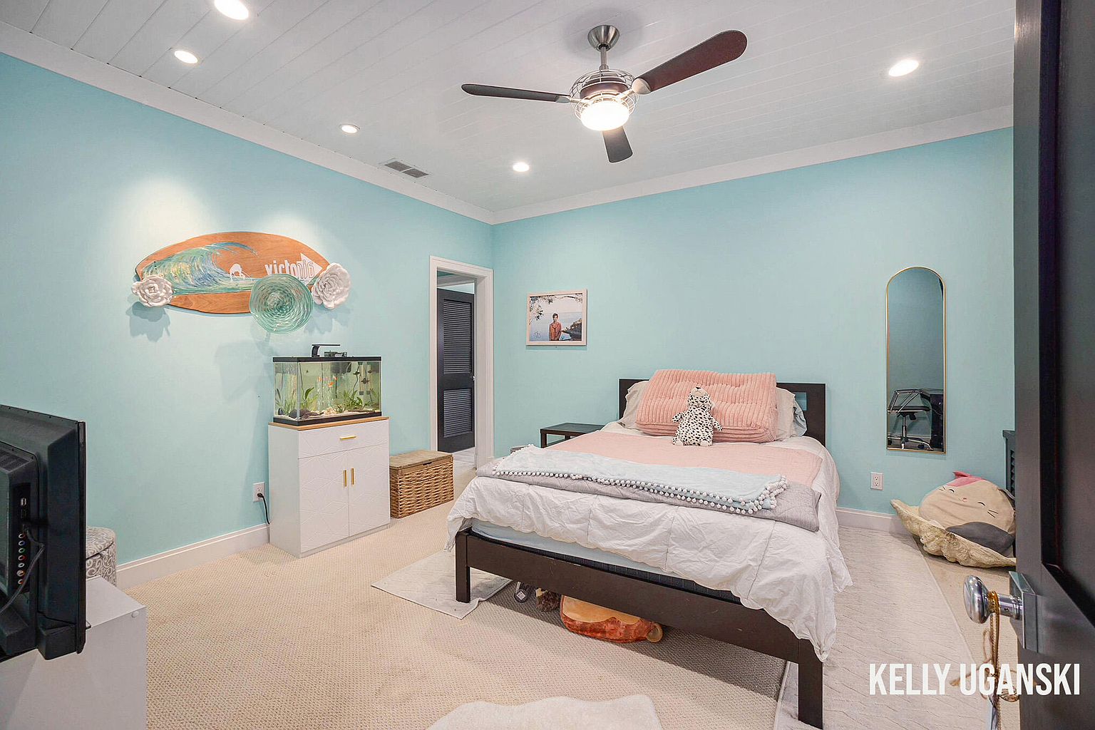 This is a cozy bedroom featuring light blue walls, a white ceiling with recessed lighting, and a ceiling fan. The room is furnished with a dark wood bed, pink bedding, and decorative items such as a surfboard wall hanging and an aquarium. The overall impression is a comfortable and inviting space.