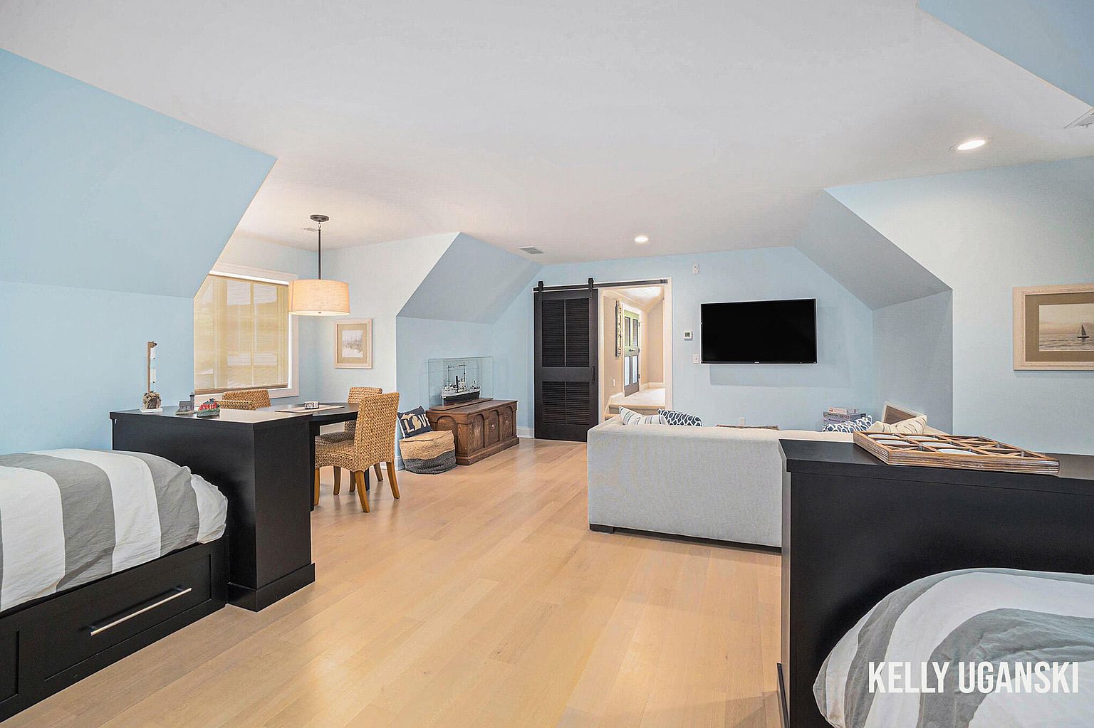 This is a bright and airy guest bedroom featuring light blue walls and light wood flooring. The room includes two beds separated by a dark wood desk, a seating area with a light gray sofa, and a dark wood sliding barn door. The room has a cozy and inviting atmosphere, perfect for guests.