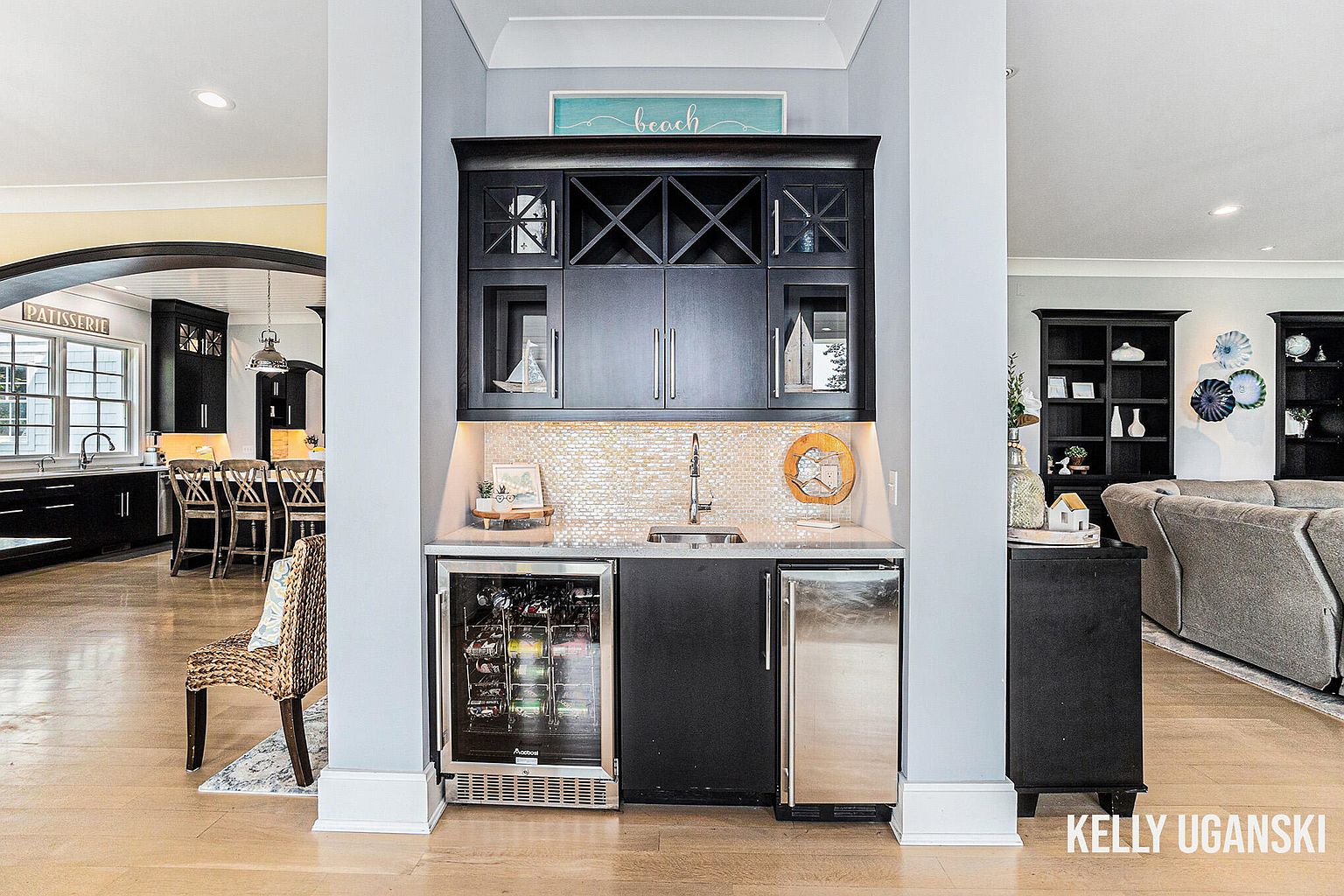 This image showcases a stylish wine cellar or bar area, featuring dark cabinetry with wine racks and glass-fronted display cabinets. A countertop with a sink and mosaic backsplash adds functionality, while a wine refrigerator and beverage cooler provide storage. The space is well-lit and appears to be integrated into the home's design, offering a sophisticated touch.