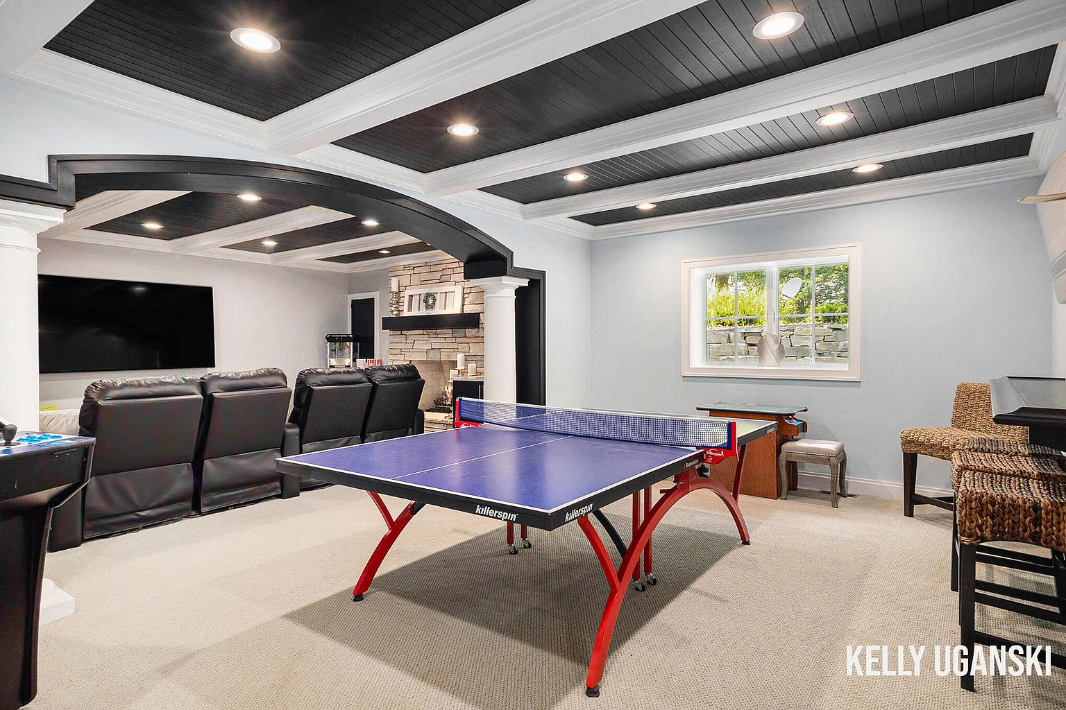 This is a well-appointed basement featuring a recreation area with a ping pong table, a comfortable seating area with leather recliners, and a stone fireplace. The ceiling has a unique coffered design with recessed lighting, and the walls are painted in a light, neutral color. The space is designed for entertainment and relaxation.