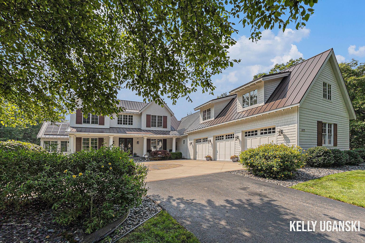 This is a front exterior view of a two-story house with a three-car garage. The house has white siding, a dark metal roof, and dark shutters. The driveway is paved and leads to a circular parking area in front of the house. Landscaping includes bushes and grass.