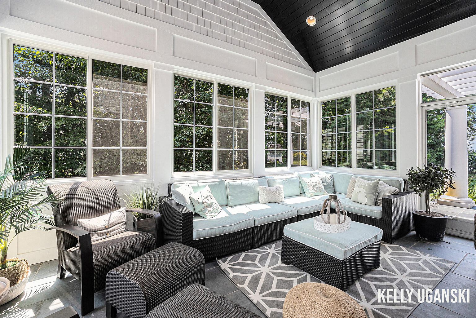 This sunroom features a black wicker sectional sofa with light blue cushions and decorative pillows, arranged around a matching ottoman and a woven pouf on a gray geometric rug. Large windows with white frames offer ample natural light and views of the surrounding greenery, while the black ceiling adds a touch of modern contrast. The space is furnished with additional seating and potted plants, creating a cozy and inviting atmosphere.