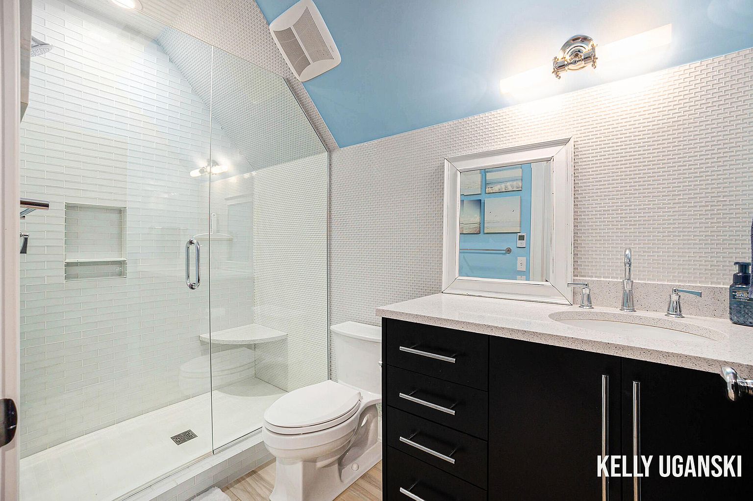 This is a well-lit bathroom featuring a glass-enclosed shower with white subway tile and a built-in seat. The vanity has a dark cabinet with a light countertop, a modern sink, and chrome fixtures. The walls are covered in a textured white tile, and the ceiling is painted a light blue, creating a clean and contemporary aesthetic.