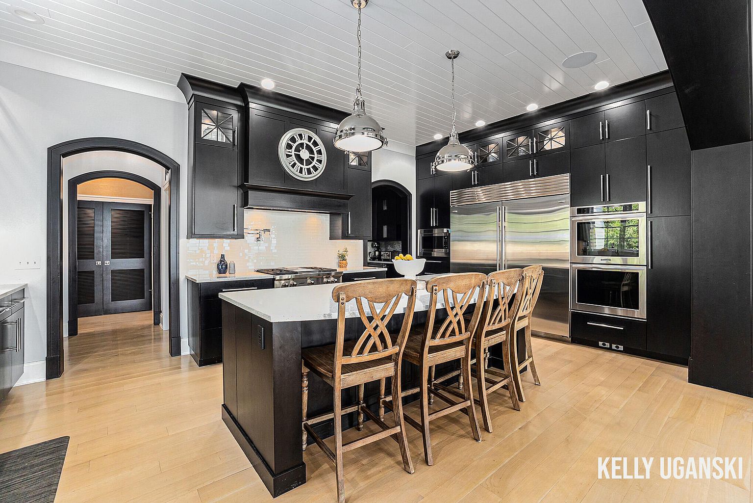 This is a well-lit kitchen featuring dark cabinetry, stainless steel appliances, and a large island with a white countertop. The kitchen has a modern and elegant style, with pendant lighting and a decorative clock adding to the ambiance. The perspective is from a wide angle, showcasing the entire kitchen space and its layout.
