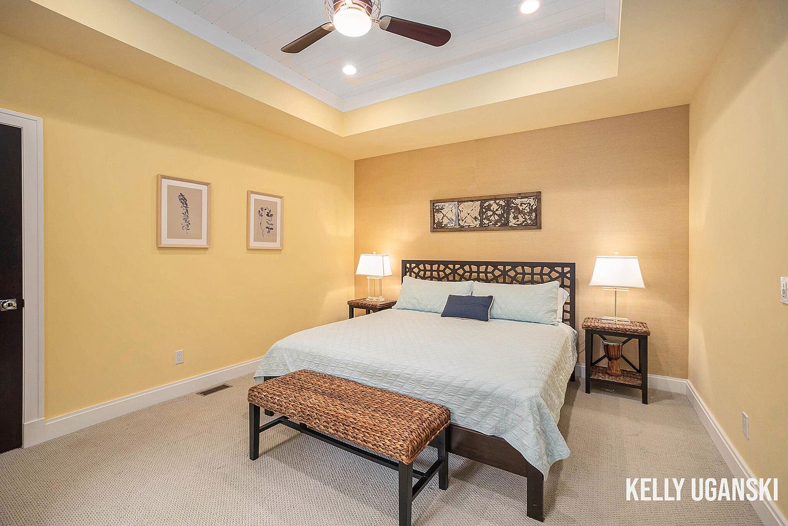 This is a cozy primary bedroom featuring a queen-sized bed with a decorative headboard and a light blue comforter. The room is painted in warm yellow tones, with one wall featuring a textured wallpaper. Two framed botanical prints hang on the wall above the bed, and a woven bench sits at the foot of the bed. Two bedside tables with lamps provide soft lighting, and a ceiling fan adds to the comfort of the room.