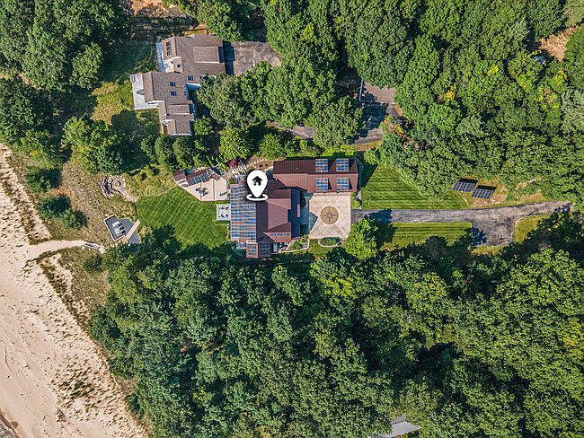 This aerial shot showcases a luxurious property nestled amidst lush greenery, offering a sense of privacy and seclusion. The house features a unique architectural design with a combination of modern and rustic elements, including a distinctive circular patio. The property also includes solar panels, a well-maintained lawn, and direct access to a sandy beach, enhancing its appeal as a high-end real estate offering.