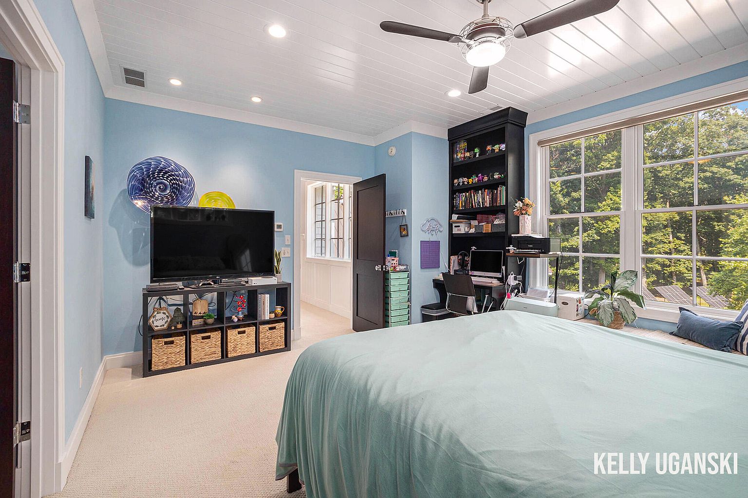 This is a cozy bedroom featuring light blue walls and a white paneled ceiling with a ceiling fan. The room includes a TV on a storage unit, a desk area with a black bookshelf, and a large window offering natural light and a view of the outdoors. The bed is covered with a light blue blanket, creating a serene and inviting atmosphere.