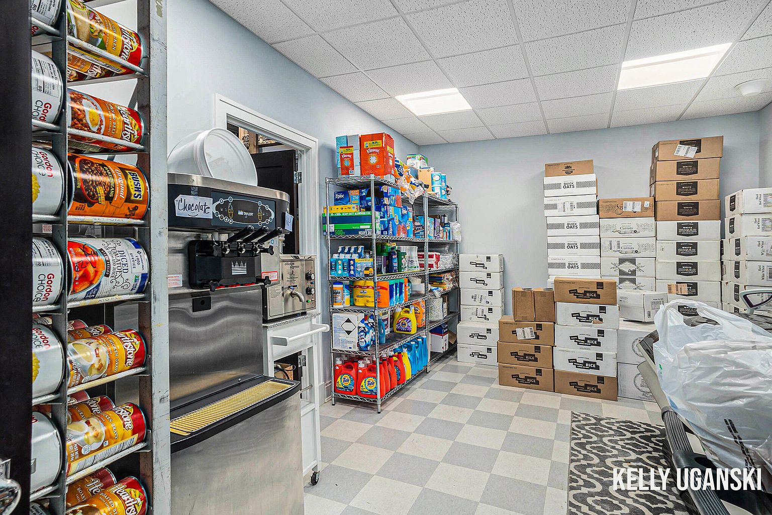 This is a well-stocked pantry featuring metal shelving units filled with canned goods, cleaning supplies, and various household items. Boxes are neatly stacked on the floor, indicating ample storage space. A commercial-grade soft serve ice cream machine is also present, adding a unique feature to the pantry.