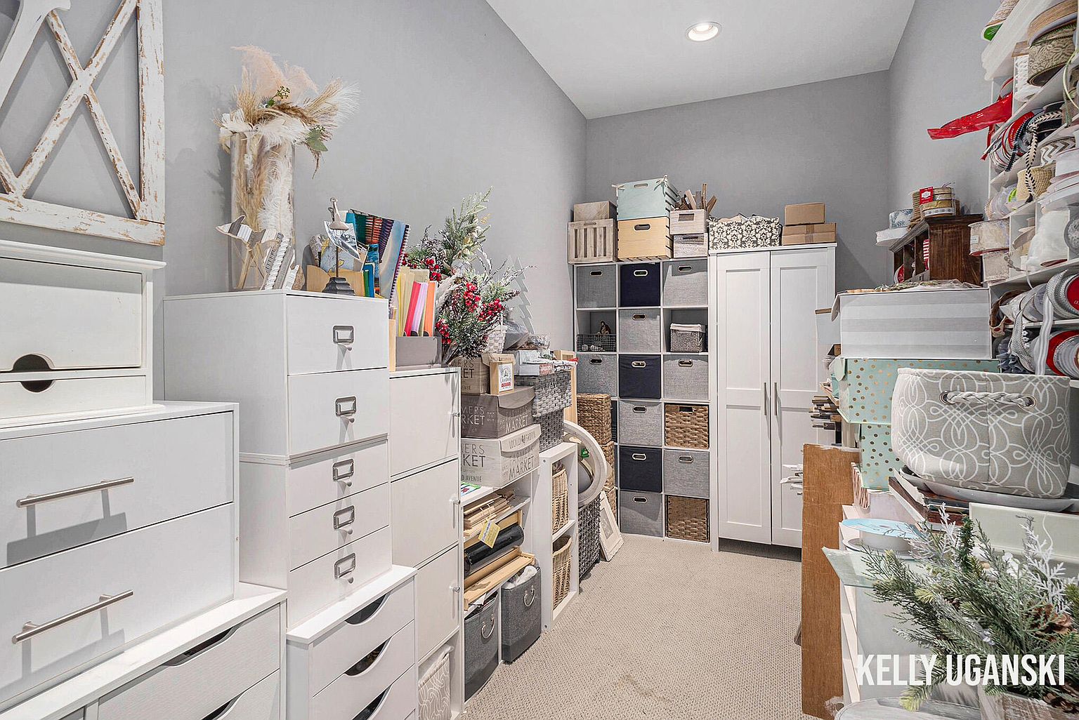 This interior shot showcases a well-organized office or study space, featuring white storage units and shelving filled with various items. The room is painted in a neutral gray tone, creating a calm and functional atmosphere. The image highlights the ample storage solutions, making it ideal for showcasing the room's organizational capabilities.