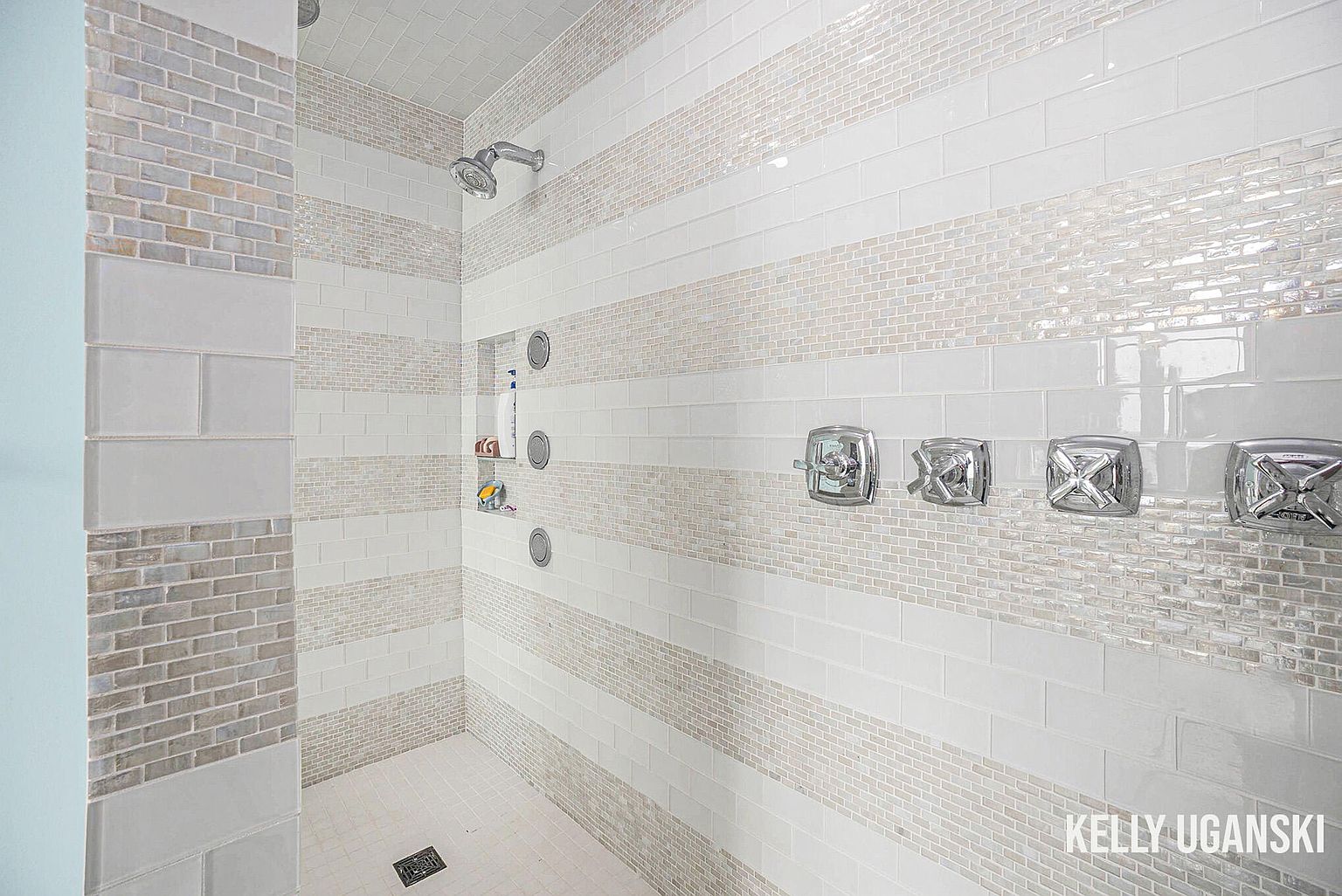 This is a well-lit primary bathroom featuring a modern shower with white and beige tiled walls arranged in horizontal stripes. The shower includes a built-in niche for toiletries, multiple shower heads, and sleek chrome fixtures. The overall impression is clean, contemporary, and luxurious.