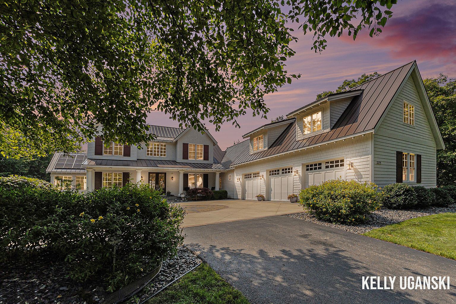 This is a front exterior view of a two-story home with a three-car garage. The house features light-colored siding, dark metal roofing, and contrasting shutters. A well-maintained lawn and landscaping enhance the property's curb appeal, and a circular driveway adds to the grandeur.