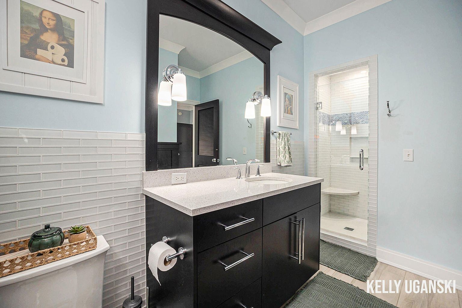 This is a well-lit bathroom featuring a dark vanity with a white countertop and chrome fixtures. A large mirror hangs above the vanity, reflecting the light and the opposite wall. The walls are painted a light blue, and the floor is a light wood, creating a clean and inviting space.