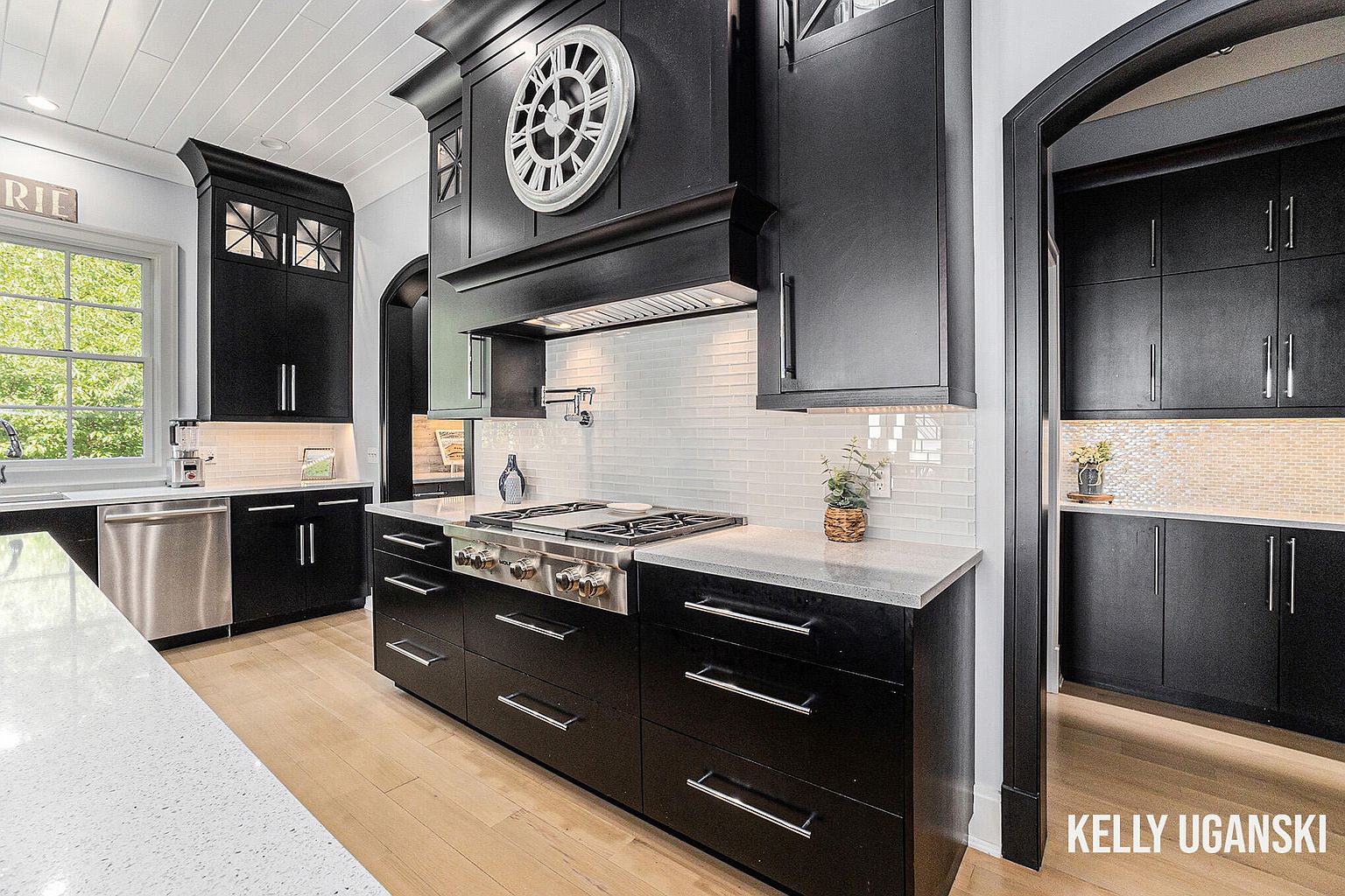 This is a modern kitchen with black cabinetry, stainless steel appliances, and light-colored countertops. The kitchen features a large range with a custom hood and a decorative clock above. The flooring is light hardwood, and the overall impression is one of sophisticated elegance.