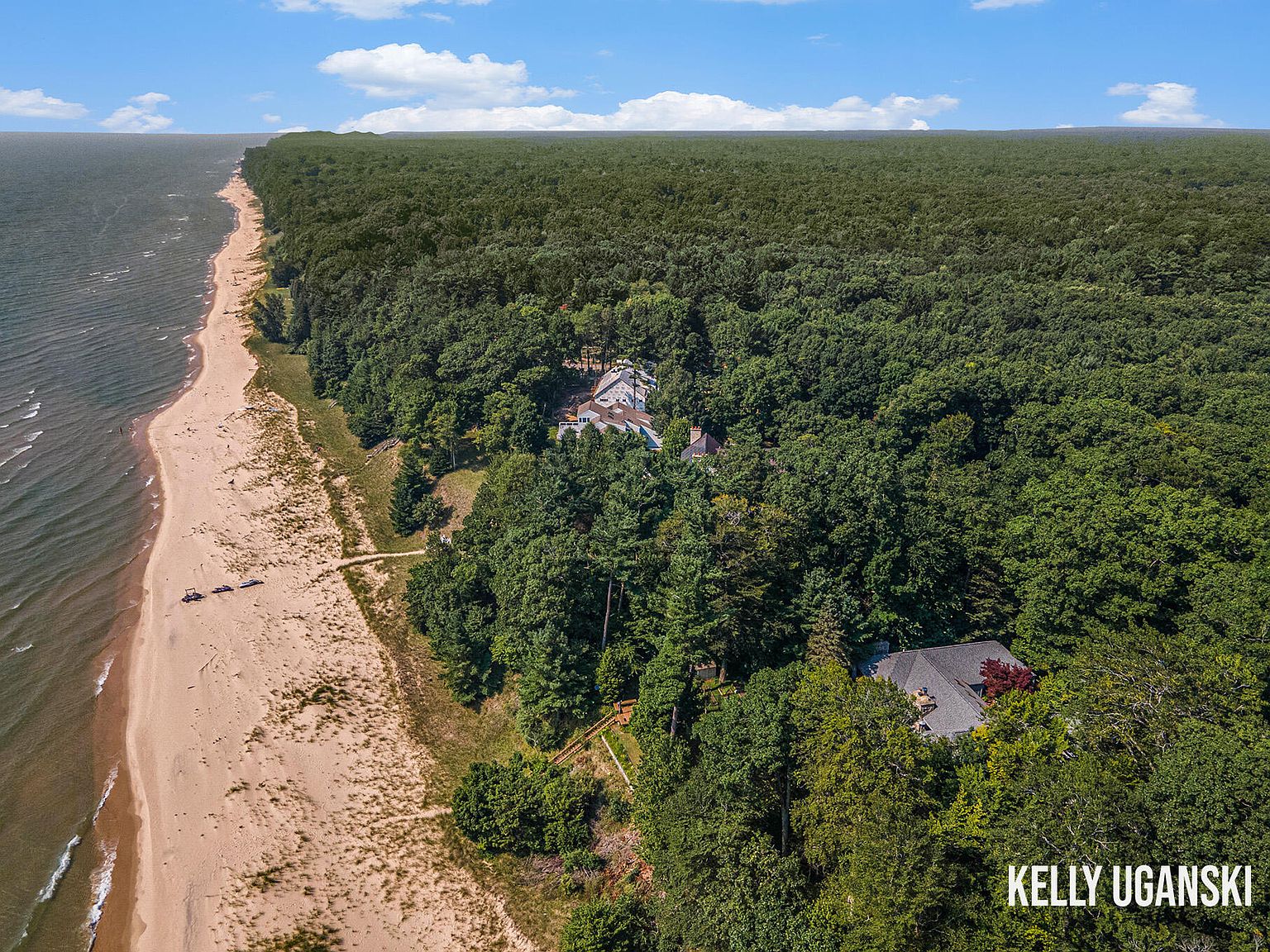 This aerial shot showcases a stunning property nestled between a lush forest and a pristine beach. The expansive sandy beach stretches along the coastline, meeting the clear waters of the lake. The property features a large house partially hidden among the trees, offering both privacy and scenic views.