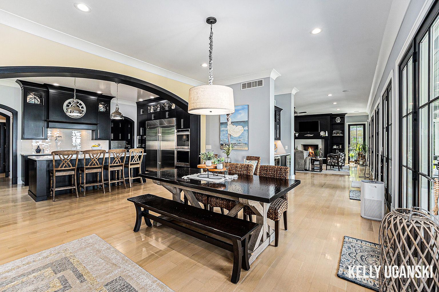 This is an interior shot of a dining area adjacent to a kitchen and living room. The dining area features a large dark wood table with a bench and woven chairs, illuminated by a pendant light. The kitchen has dark cabinetry and stainless steel appliances, while the living room has a fireplace and comfortable seating. The overall impression is of a spacious and well-appointed home with an open floor plan.