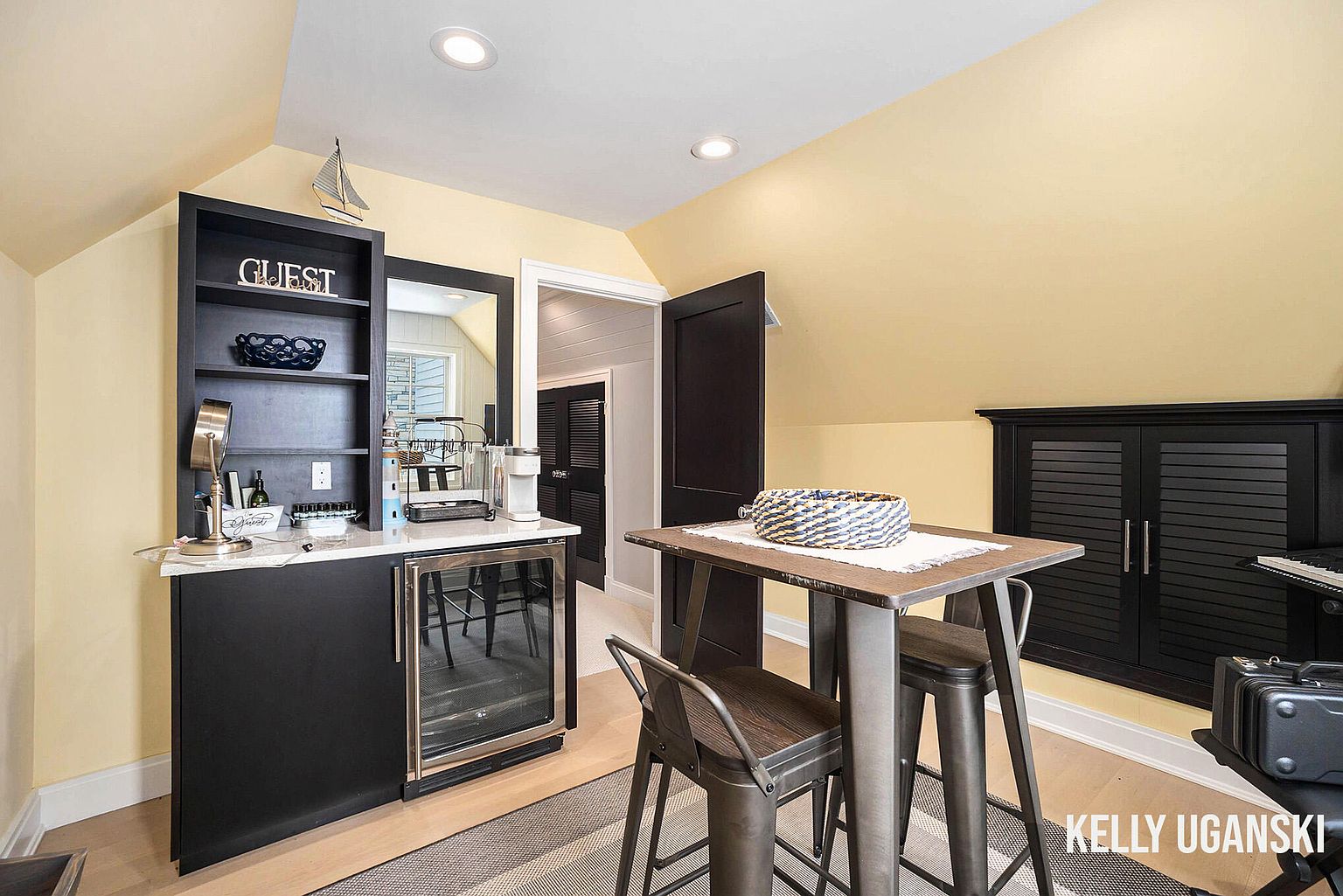 This interior shot showcases a cozy guest area, possibly in an attic or bonus room, featuring a built-in bar with a wine fridge and shelving. A small table with bar stools provides a seating area, and a dark cabinet is visible in the background. The room is painted in a warm yellow tone, creating an inviting atmosphere.