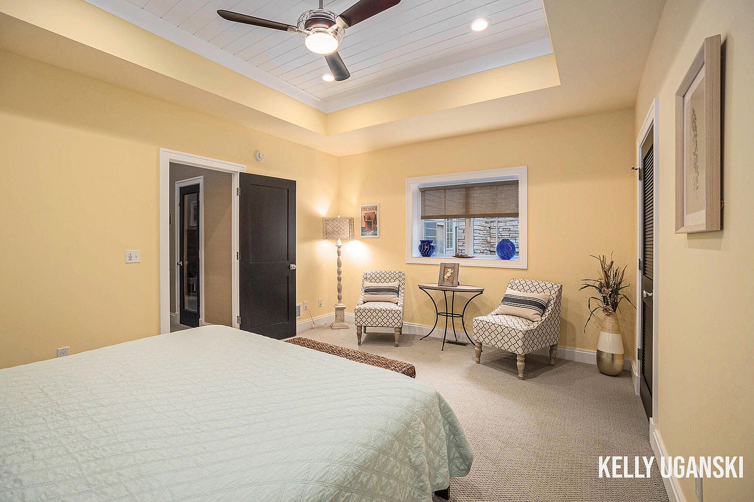 This is a primary bedroom featuring yellow walls, a tray ceiling with a ceiling fan, and carpeted floors. The room includes a bed with a light-colored quilt, two armchairs flanking a small table near a window, and a dark-colored door leading to another room. The overall impression is a cozy and well-lit space.