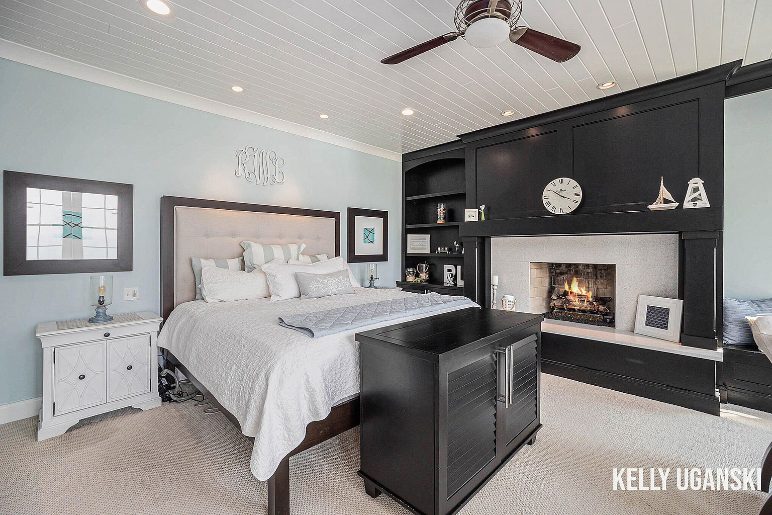This primary bedroom features a cozy fireplace with a dark wood surround and built-in shelving, creating a focal point. The bed is adorned with white linens and decorative pillows, complemented by a light blue accent wall. A white nightstand and a dark wood cabinet add to the room's functionality and style, while the overall impression is one of comfort and relaxation.