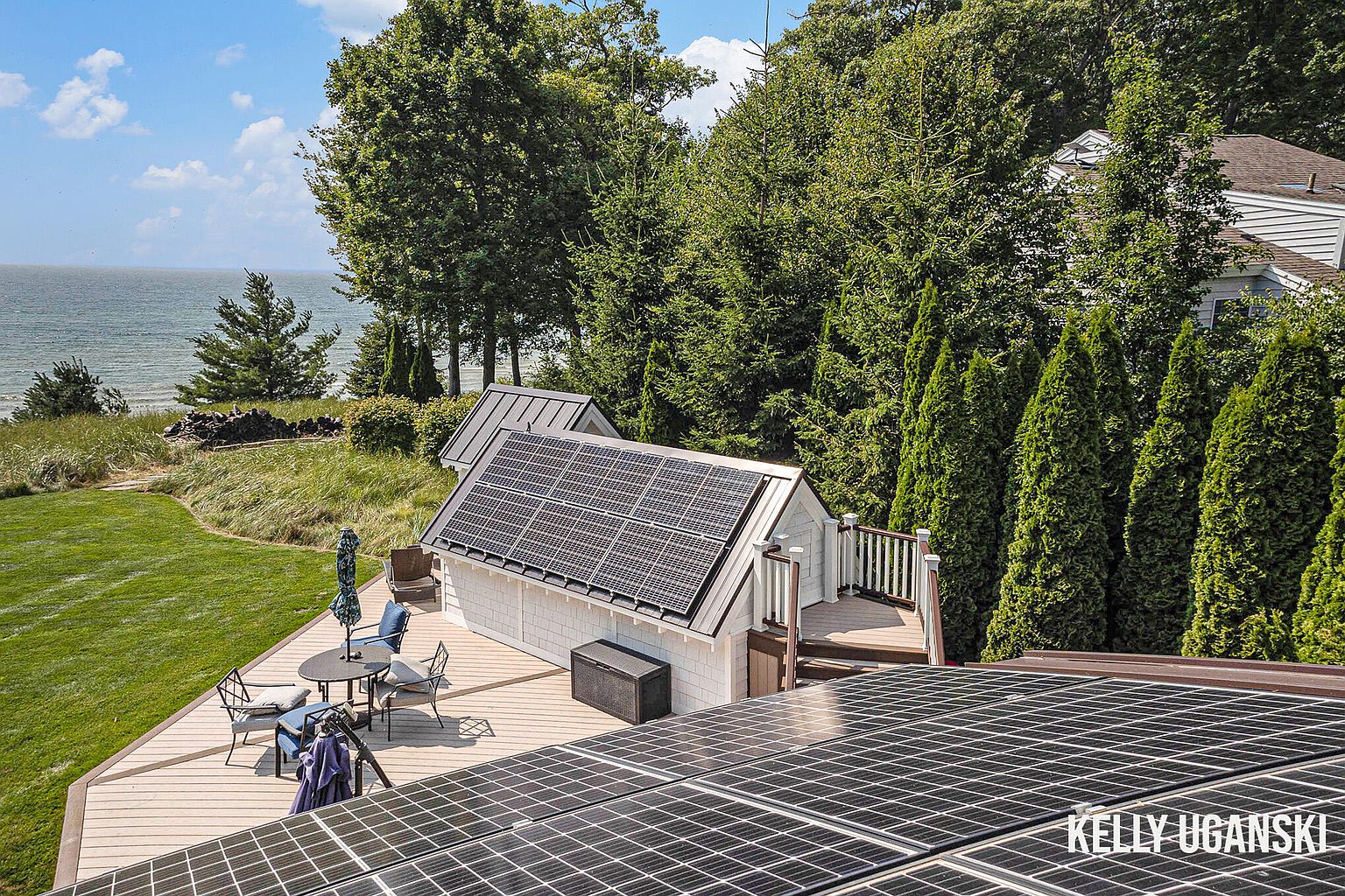 This aerial view showcases a property with a focus on its outdoor living spaces and solar panel installations. A wooden deck features a patio set, suggesting an area for relaxation and entertainment. The presence of solar panels on the roofs of the buildings highlights the property's commitment to energy efficiency and sustainability, while the surrounding trees and a glimpse of the water add to the scenic appeal.