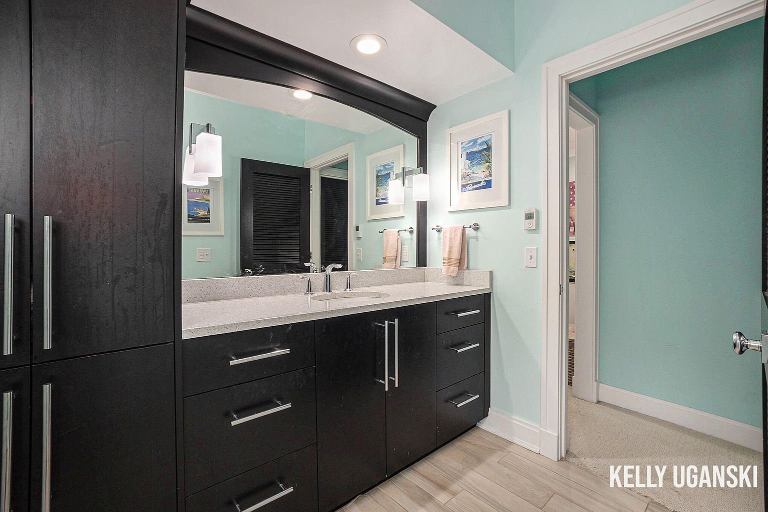 This is a bathroom featuring a dark vanity with a light countertop and silver hardware. A large mirror with a dark frame hangs above the vanity, reflecting the light blue walls and artwork. The flooring appears to be light-colored wood-look tile, and a doorway leads to another room, creating an open and airy feel.