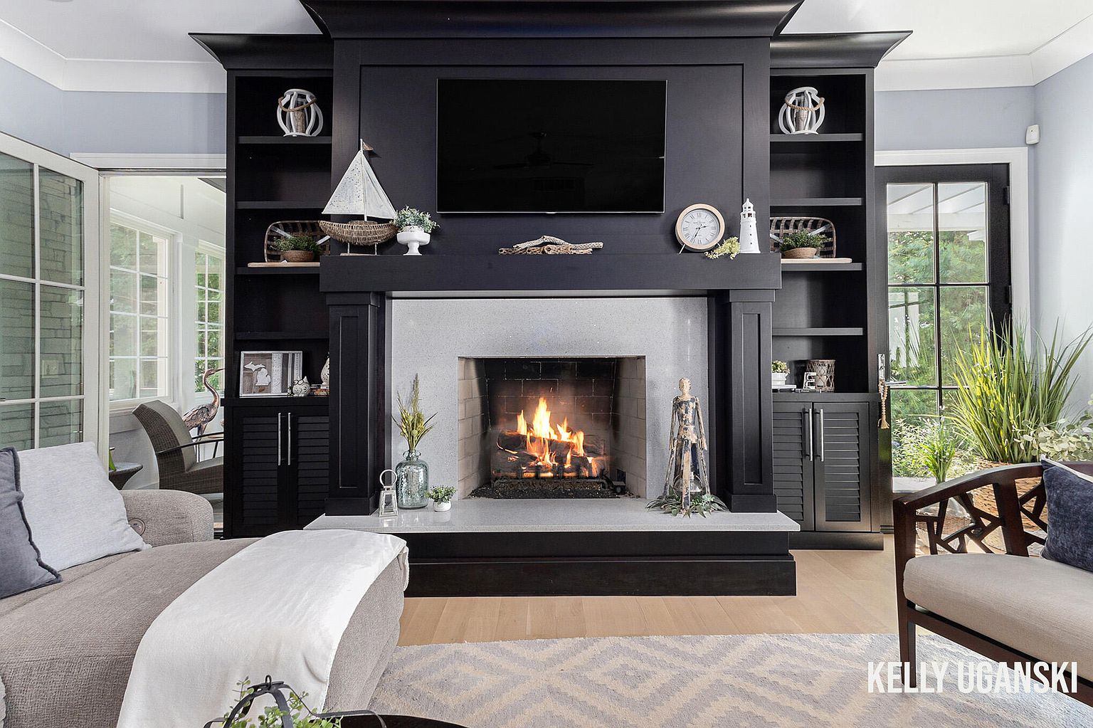 This is an interior shot of a living room featuring a fireplace with a large black built-in unit surrounding it. The fireplace is lit, and decorative items are placed on the mantel and shelves. The room has a cozy and inviting atmosphere, with a neutral color palette and comfortable seating.