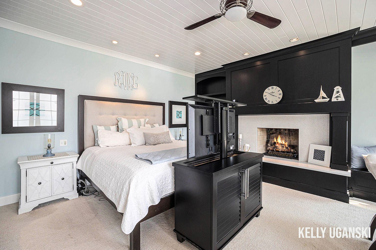 This primary bedroom features a neutral color palette with light blue walls and white trim. A large bed with a tufted headboard is the focal point, complemented by a dark wood entertainment center with a fireplace. The room is well-lit with recessed lighting and a ceiling fan, creating a cozy and inviting atmosphere.