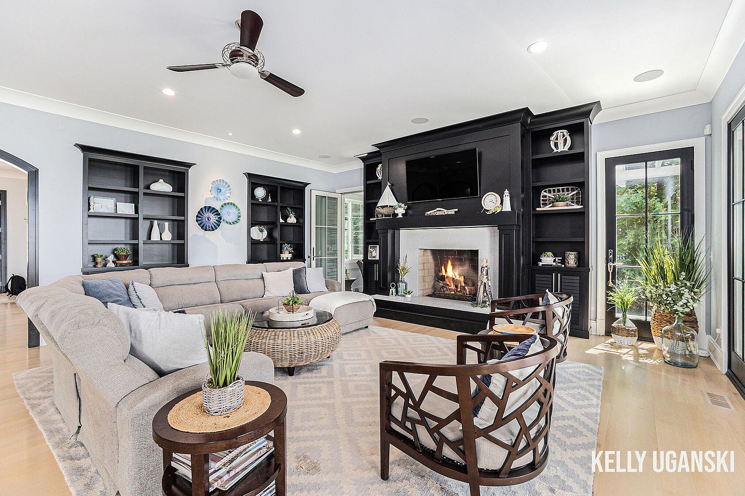 This is a well-lit living room featuring a large sectional sofa, a fireplace with a black surround and built-in shelving, and two accent chairs. The room is decorated in a neutral color palette with pops of blue and green, creating a cozy and inviting atmosphere. The flooring is light wood, and a patterned area rug anchors the seating arrangement.