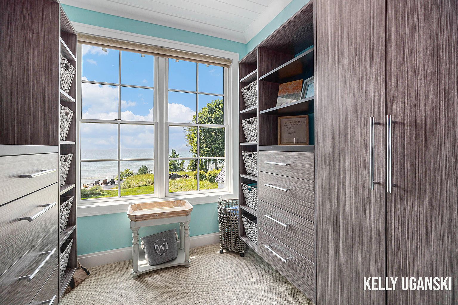 This is a well-organized walk-in closet featuring custom shelving and drawers in a modern style. The closet has a large window with a view of the water, bringing natural light into the space. The color palette is neutral, with light blue walls and light carpet, creating a serene and luxurious atmosphere.