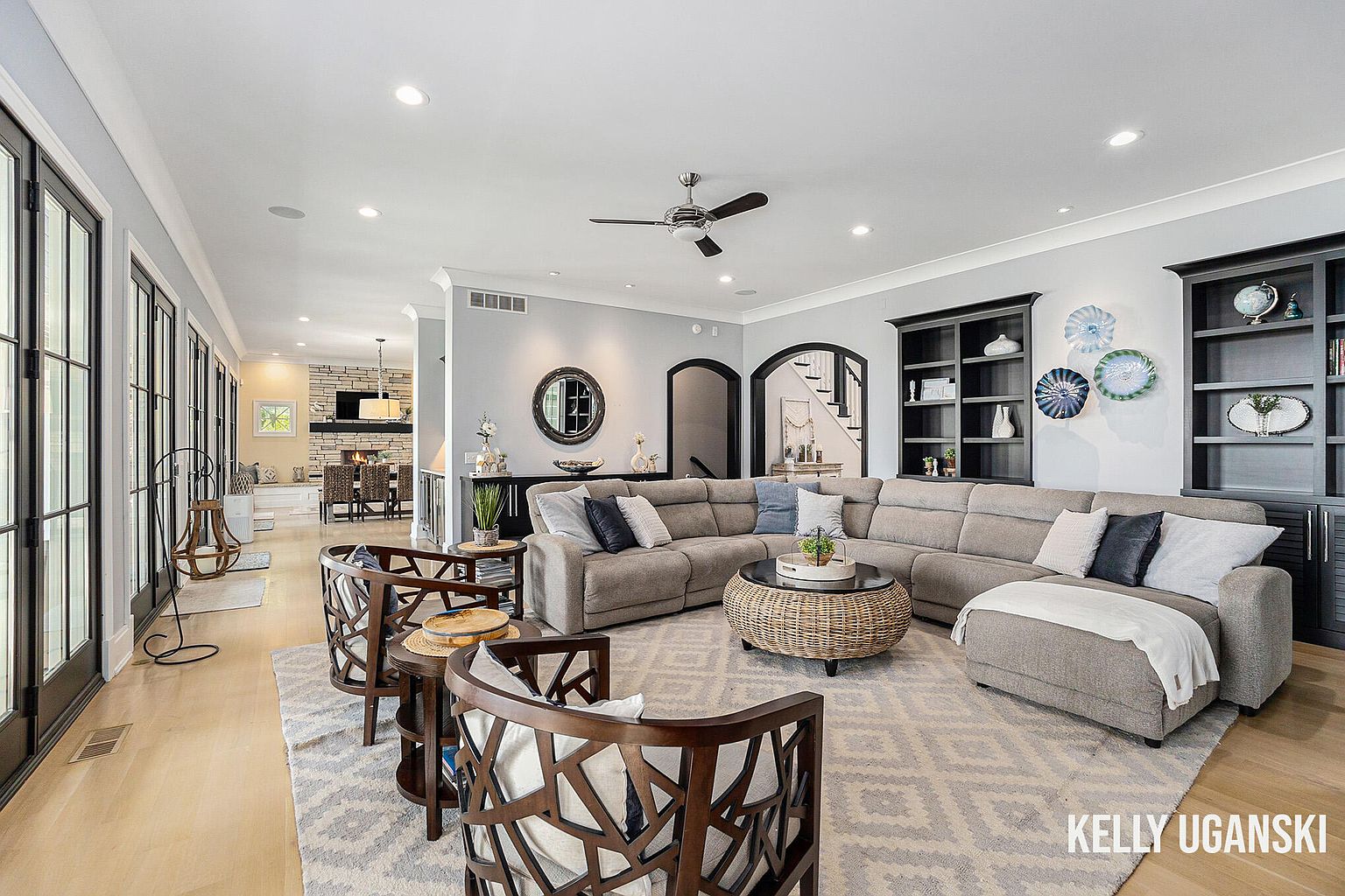 This is an interior shot of a living room featuring a large sectional sofa, wooden chairs, and a round coffee table. The room has hardwood floors, a neutral color palette, and built-in bookshelves. The space is well-lit with recessed lighting and natural light from the adjacent room, creating a comfortable and inviting atmosphere.