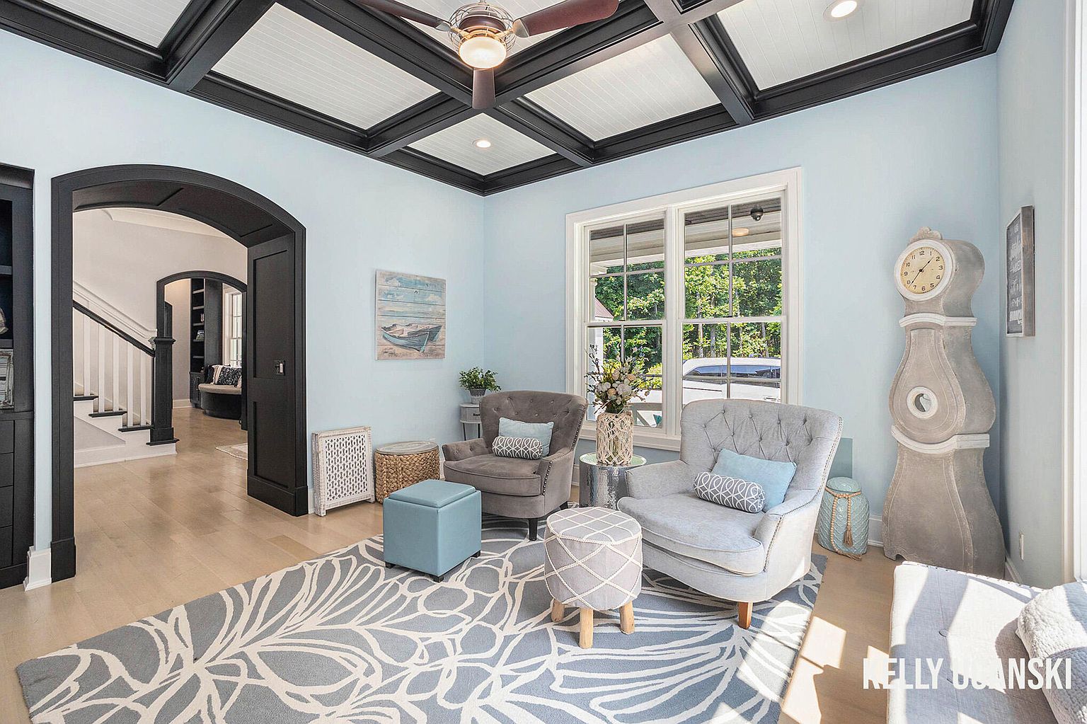 This is an interior shot of a living room featuring light blue walls, a coffered ceiling with dark beams, and light wood flooring. The room is furnished with two armchairs, ottomans, and a large area rug with a floral pattern. A grandfather clock stands near the window, and an arched doorway leads to another room, creating an inviting and stylish living space.