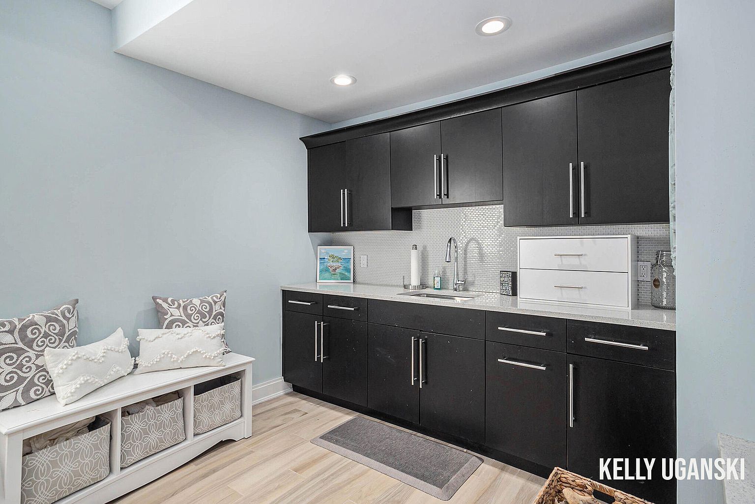 This is a well-organized laundry room featuring dark cabinetry with silver hardware, a white countertop, and a white tiled backsplash. A white bench with storage baskets and decorative pillows adds a touch of comfort and style. The room is painted in a light blue color, creating a clean and inviting space.