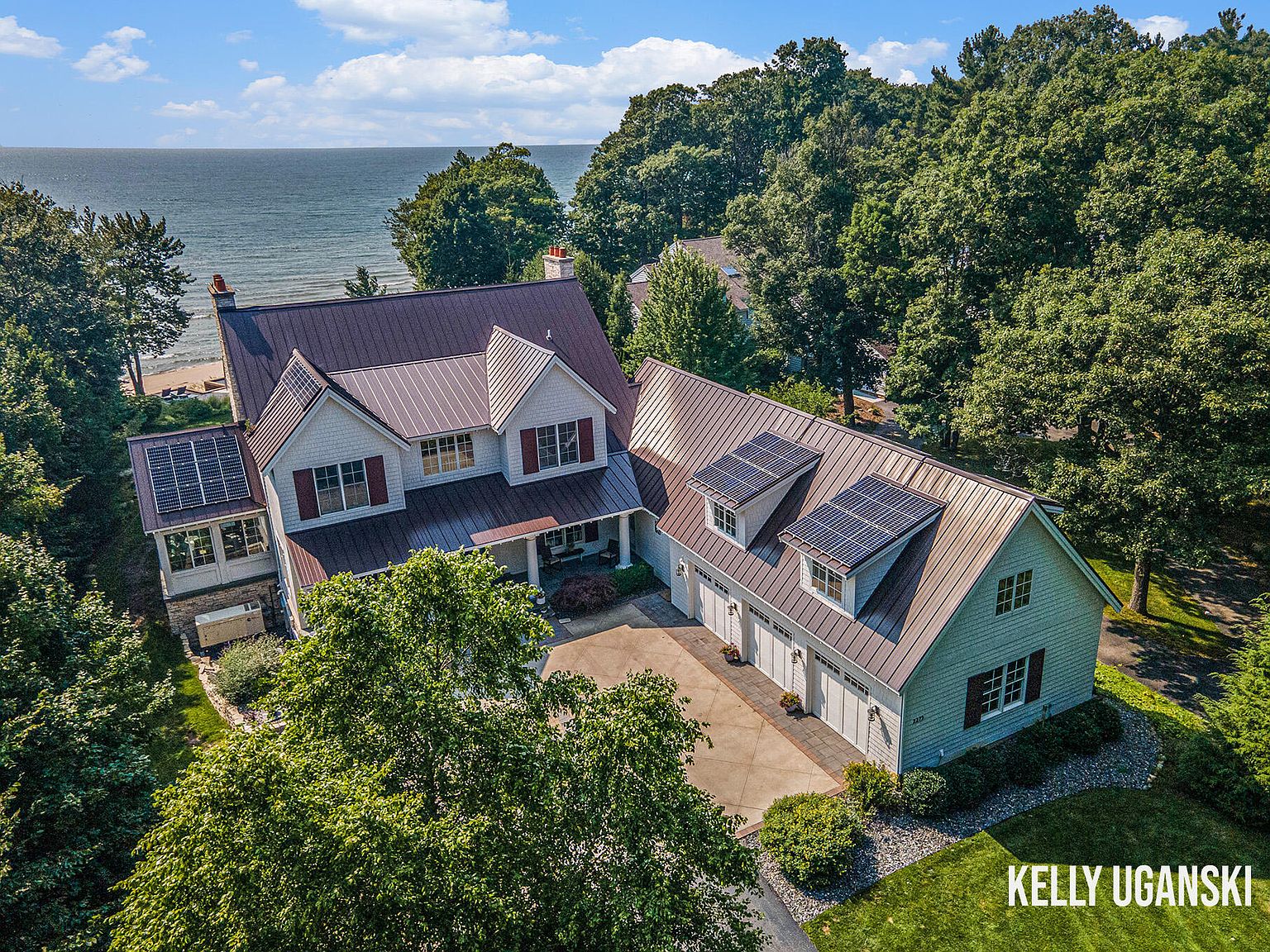 This aerial view showcases a stunning property with a main house and a detached garage, both featuring metal roofs and solar panels. The house is situated near a body of water, possibly a lake or ocean, with lush greenery surrounding the buildings. The overall impression is one of luxury, sustainability, and a desirable location.