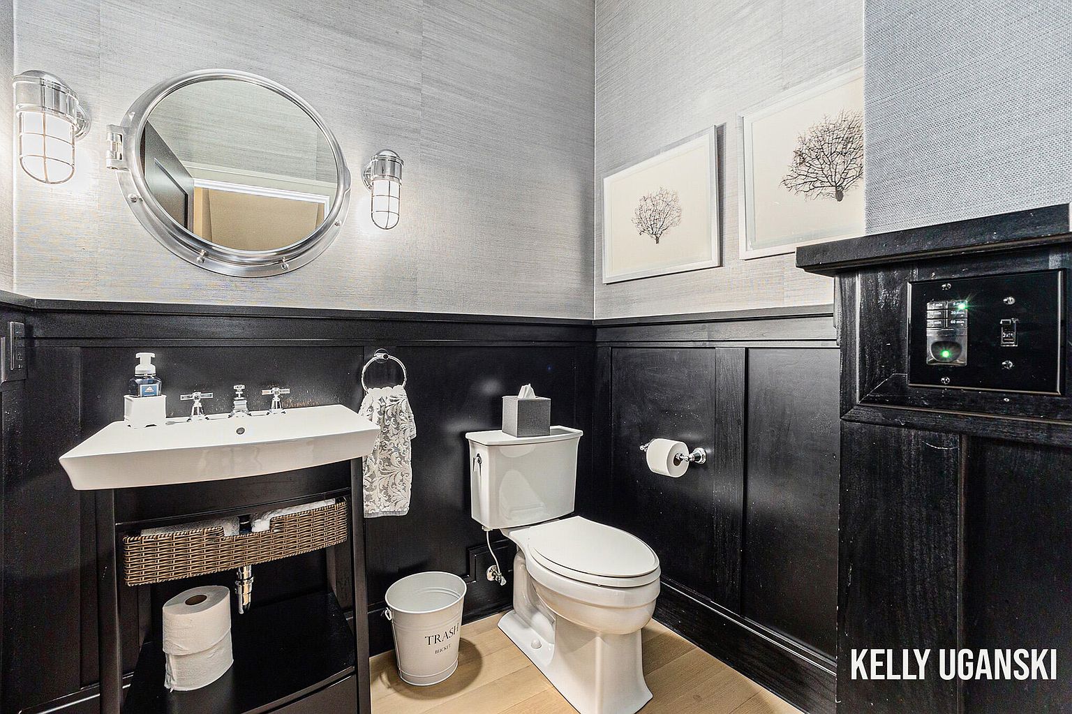 This is a stylish guest bathroom featuring a unique round mirror with nautical-inspired sconces. The walls are a textured gray above a black wainscoting. A modern sink and toilet are present, along with framed artwork, creating a sophisticated and well-appointed space.