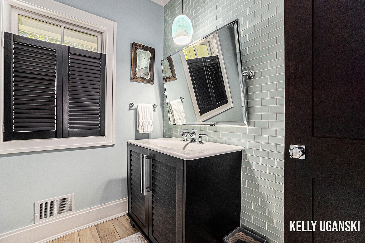 This is a well-lit guest bathroom featuring a dark vanity with a white countertop and a modern sink. The walls are painted a soft blue, complemented by light blue subway tile on one wall. A unique mirror hangs above the sink, and a window with dark shutters provides natural light, creating a clean and stylish space.