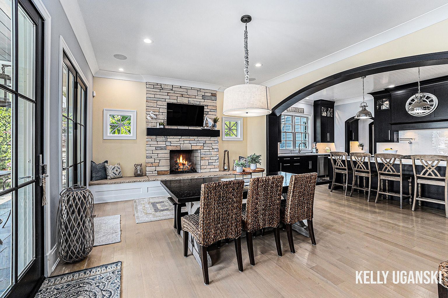 This interior shot showcases a dining area adjacent to a kitchen and living space. The dining room features a dark wood table surrounded by woven chairs, illuminated by a modern chandelier. A stone fireplace with a mounted TV adds a cozy focal point, while the open layout creates a seamless flow between the dining area and the adjacent kitchen with its dark cabinetry and bar seating.
